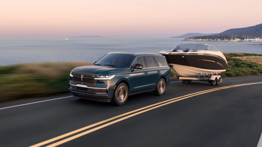 The 2025 Lincoln Navigator® Reserve in a rich green finish, towing a sleek black-and-white boat along a coastal highway at sunset.