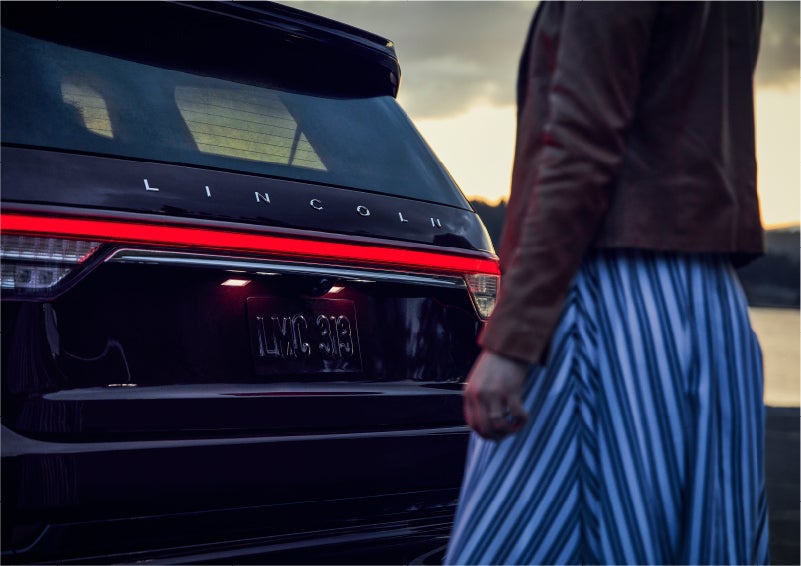 A person is shown near the rear of a 2023 Lincoln Aviator® SUV as the Lincoln Embrace illuminates the rear lights | Nick Mayer Lincoln Westlake in Westlake OH