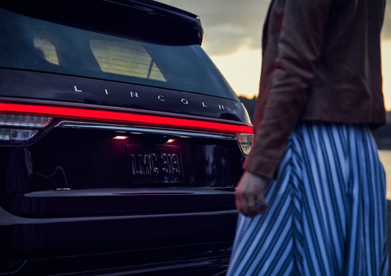 A person is shown near the rear of a 2024 Lincoln Aviator® SUV as the Lincoln Embrace illuminates the rear lights | Nick Mayer Lincoln Westlake in Westlake OH