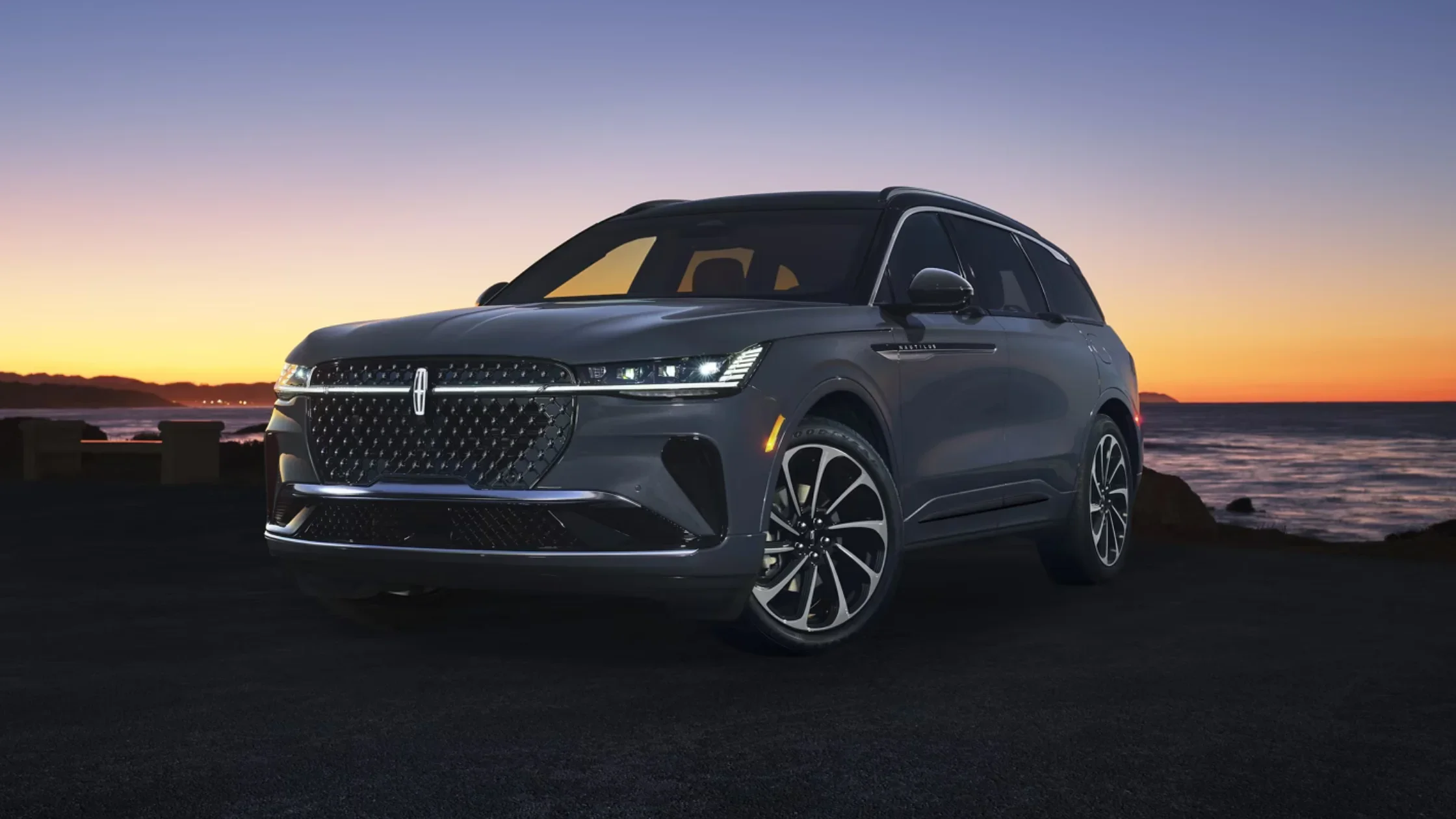 Gray 2025 Lincoln Black Label Nautilus parked near ocean at sunset with illuminated grille and coastal backdrop.