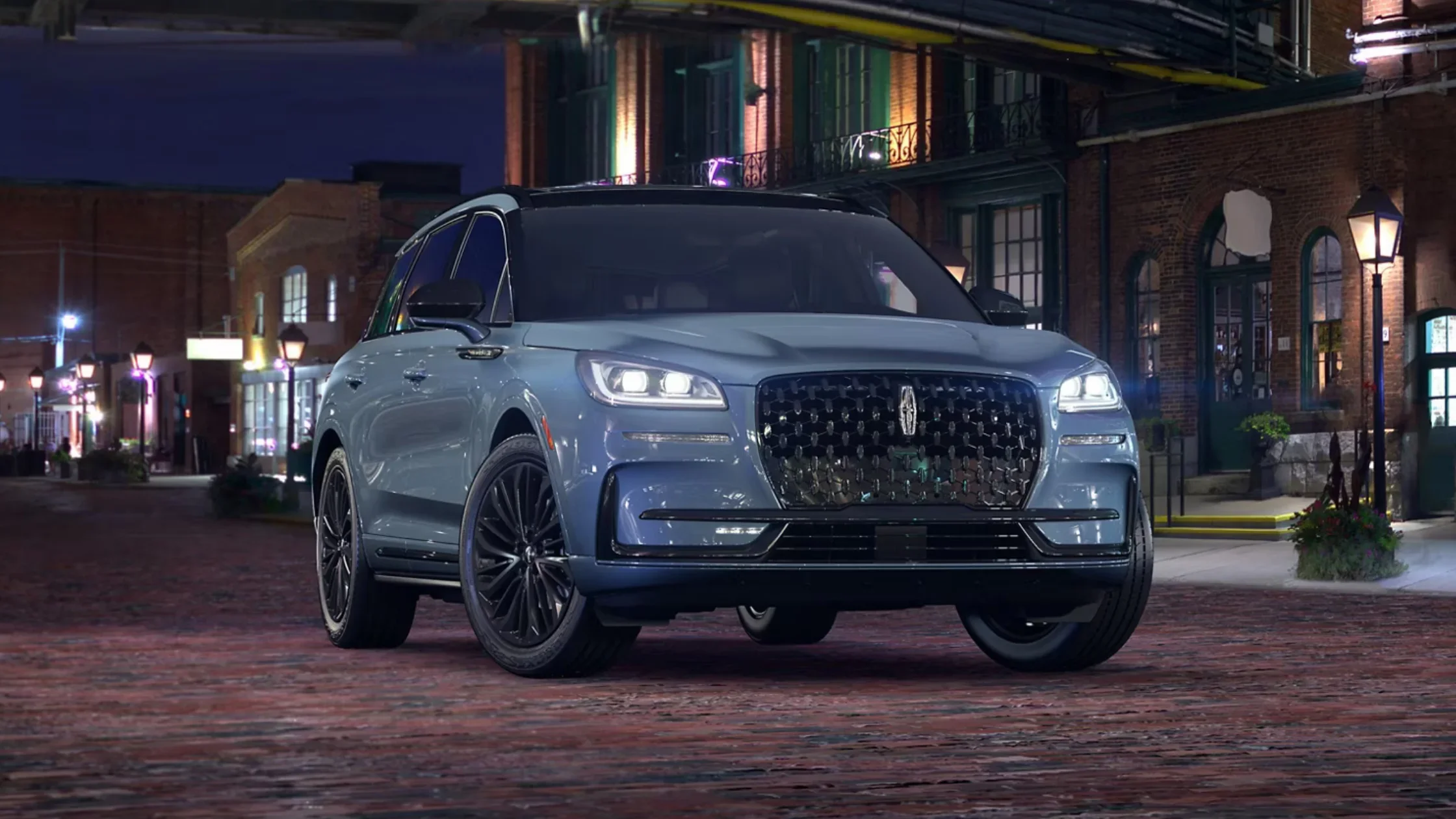 Front angle view of a 2025 Lincoln Corsair® Grand Touring in metallic blue parked on a cobblestone street at night, with LED headlights and signature grille illuminated.