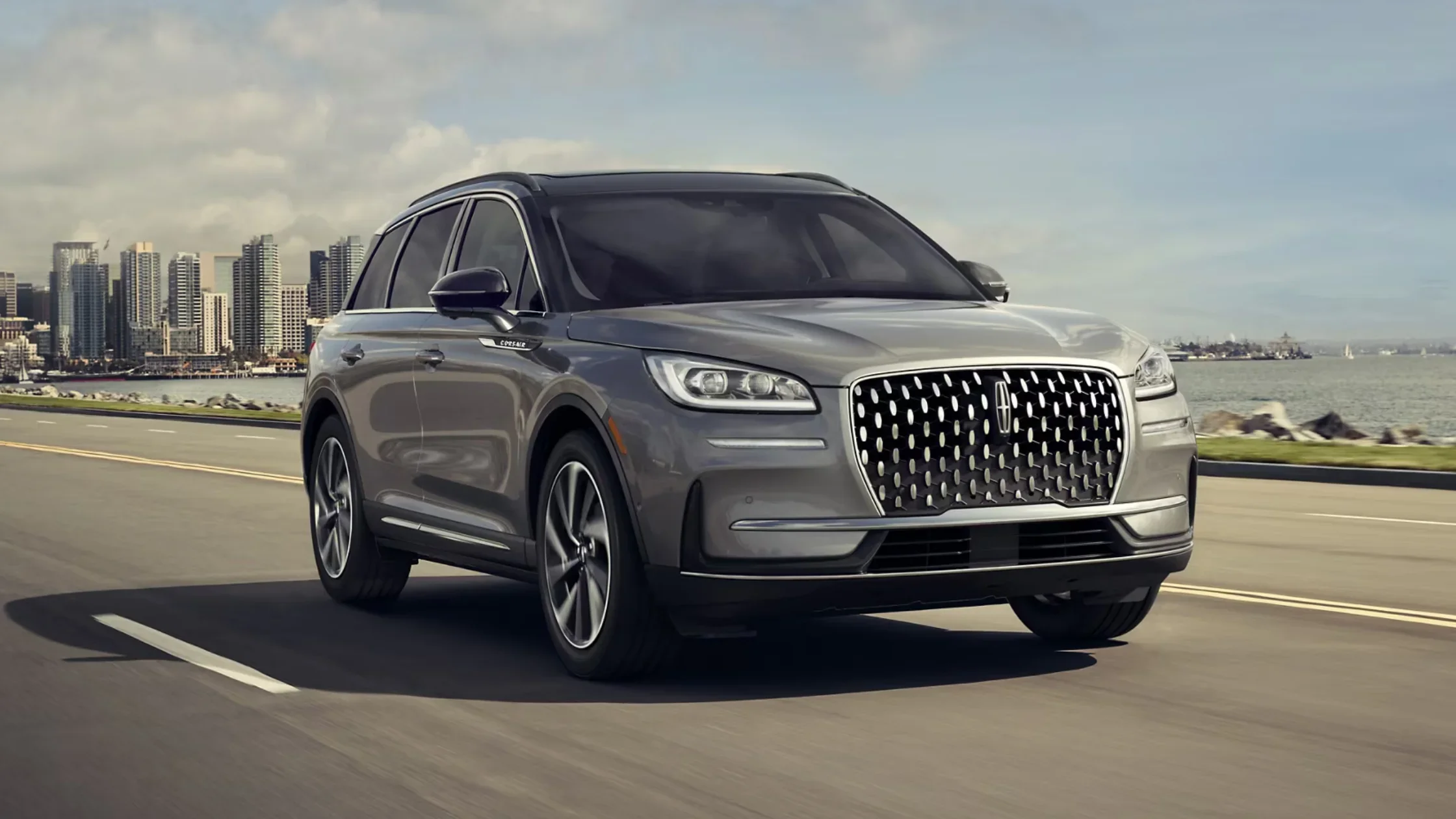 Gray 2025 Lincoln Corsair driving on highway near waterfront with a city skyline in the background.