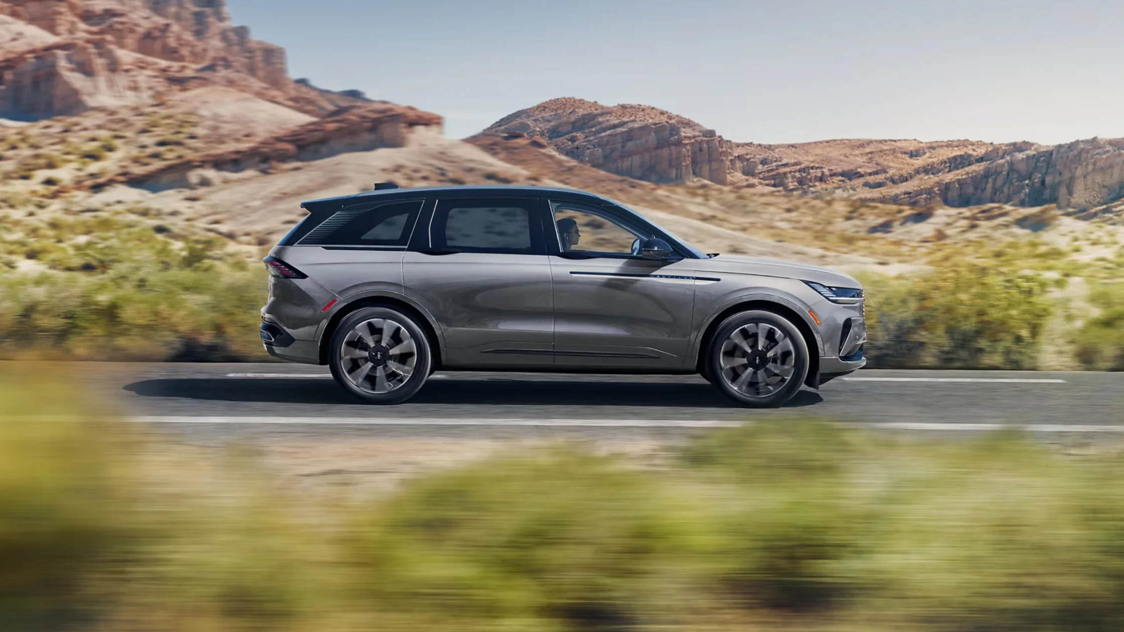 Side profile of gray 2025 Lincoln Nautilus driving through the desert with rock formations in the distance.