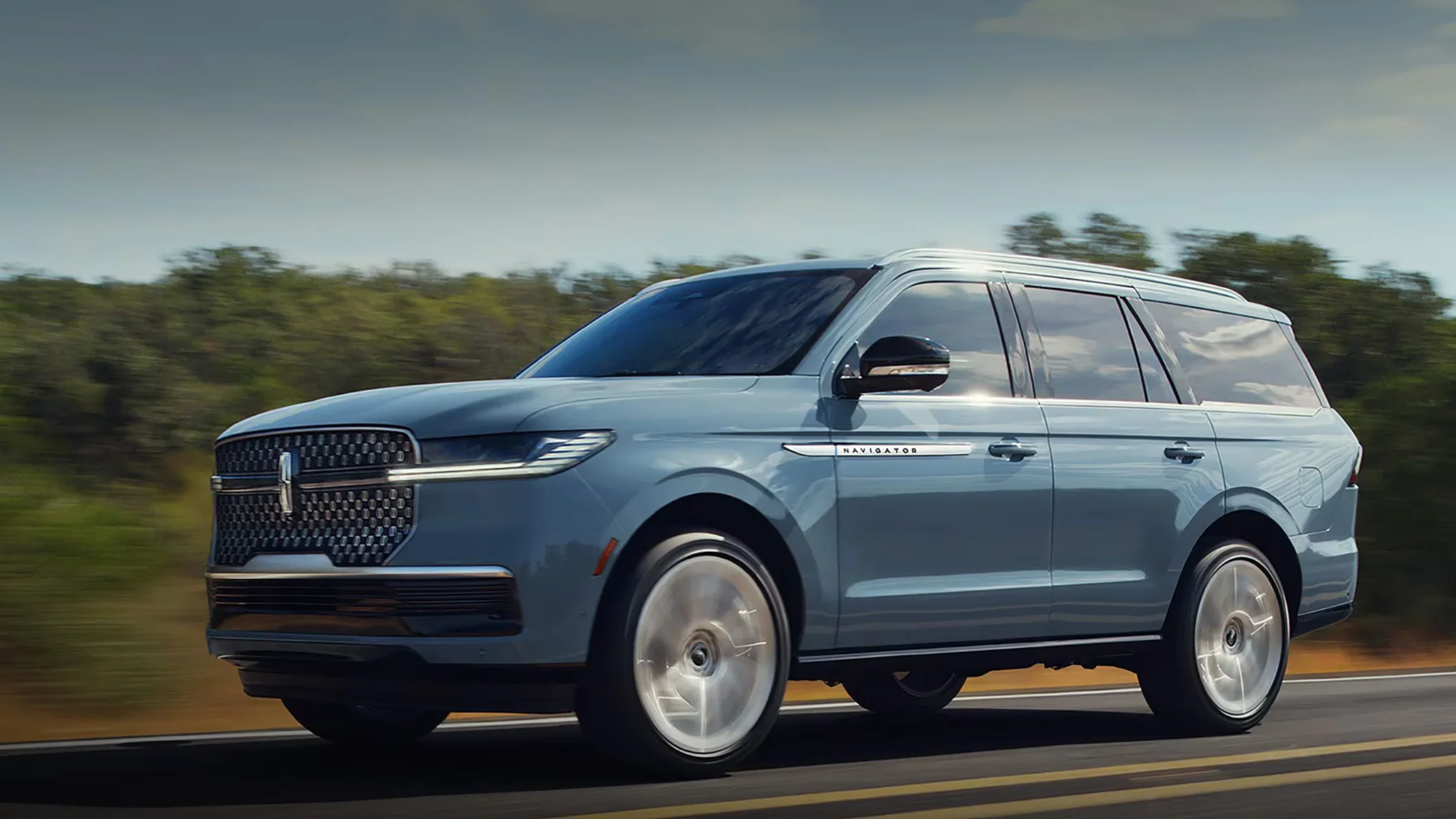 Light blue 2025 Lincoln Navigator® Reserve driving on a forest-lined road, showcasing its signature grille, sleek headlights, and bold Navigator badging.