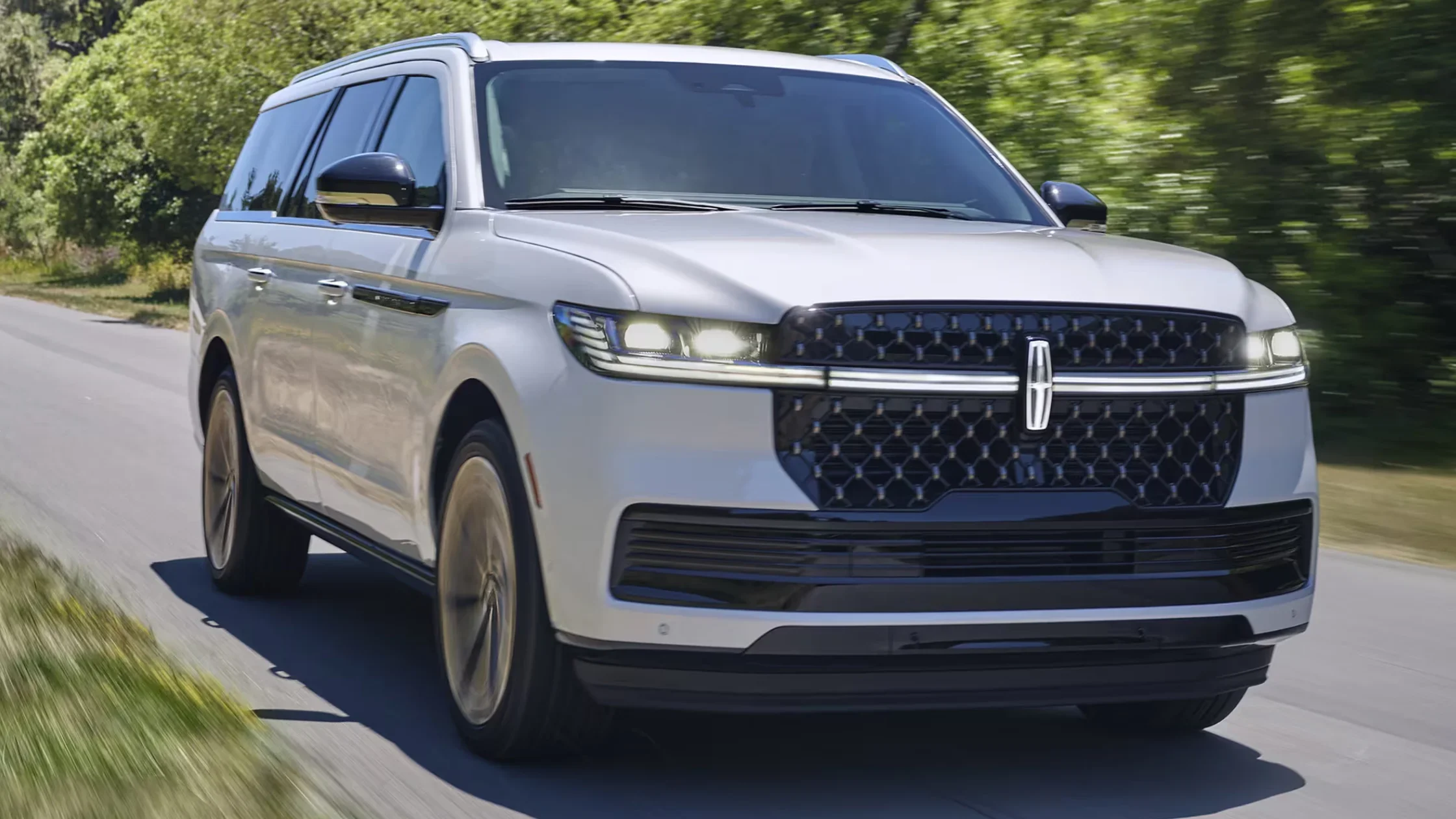 Front angle of silver 2025 Lincoln Navigator driving on shaded road with LED headlights and black grille.