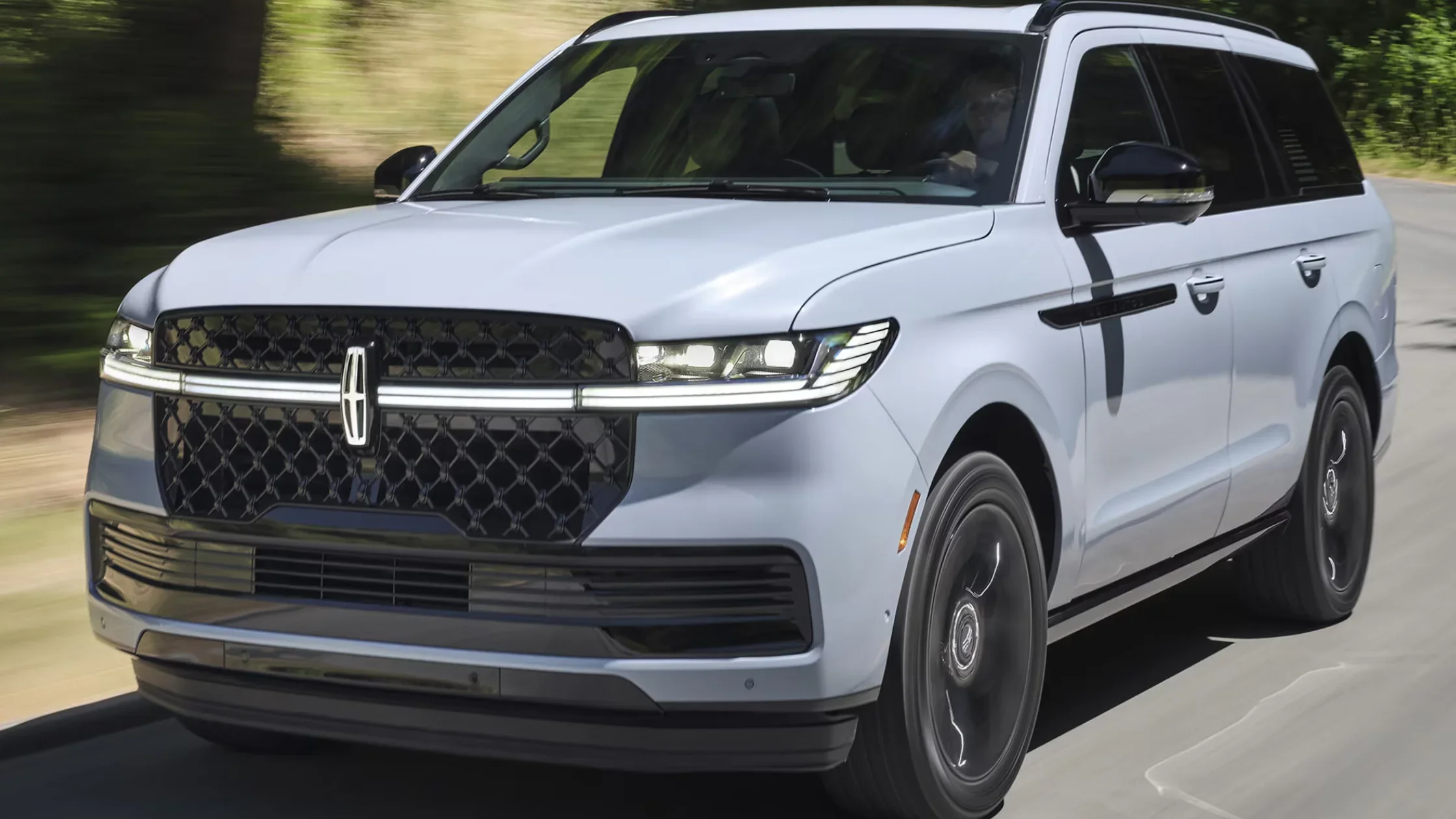 Blue 2025 Lincoln Navigator towing a boat on coastal highway, showcasing its power aWhite 2025 Lincoln Navigator driving on a forest-lined road with signature mesh grille and LED lighting.and trailering capability.