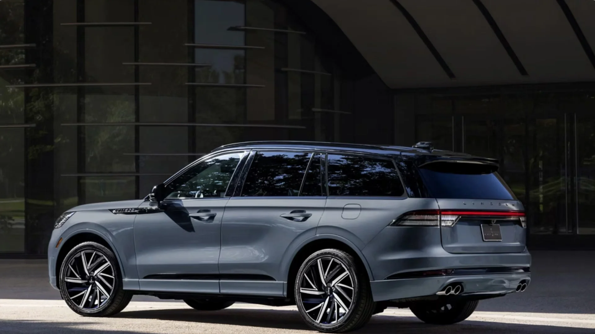 Rear three-quarter view of a 2025 Black Label Aviator® in a metallic gray finish, highlighting its sleek body lines, quad exhaust outlets, and premium wheels parked beside a modern glass building.