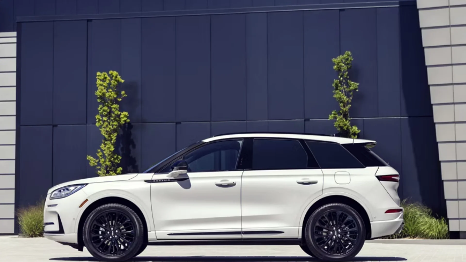 Side profile of the 2025 Corsair® Reserve in white featuring black alloy wheels against a modern building backdrop.