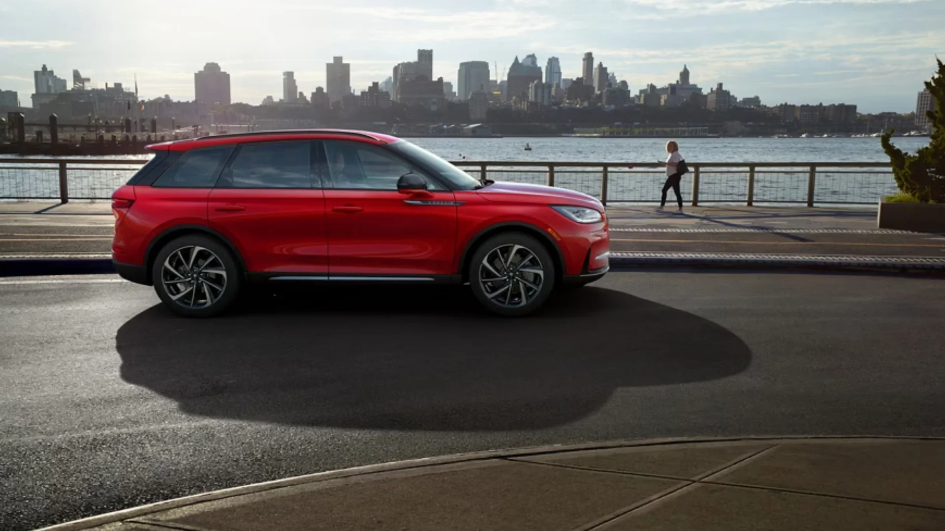 Side profile of a red 2025 Lincoln Corsair® Reserve parked near a waterfront with a city skyline in the background, showcasing its sleek exterior design and alloy wheels.
